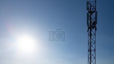 This image features a stark telecommunication tower silhouetted against a bright and clear sky, with the sun beaming directly behind it, creating a flare effect. The suns rays envelop the structure
