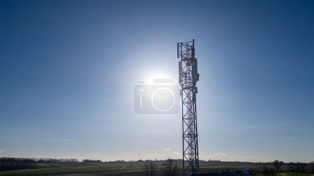 This photograph captures a solitary telecommunication tower standing tall against a bright blue sky, backlit by the suns powerful rays, which create a halo effect around the structure. The towers