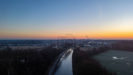 This serene image captures the calm of dawn as the days first light graces a frosty canal winding through a waking town. The gradient of the sky from the cool blues of night to the warm oranges of