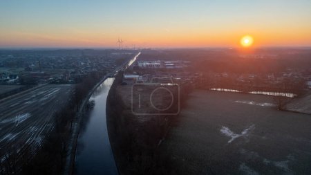 As the sun descends on the horizon, its warm glow reflects off the calm waters of the canal, which runs parallel to the rural landscape. This aerial shot captures the peaceful coexistence of nature