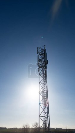 Dieses vertikale Bild zeigt eine hoch aufragende Silhouette eines Telekommunikationsturms vor dem Hintergrund eines klaren blauen Himmels, wobei die Sonne hell scheint und einen Fackeleffekt erzeugt. Der Turm