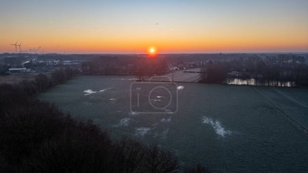 Dieses Bild verkörpert die heitere Schönheit eines kalten Morgens, an dem die Sonne über einer frostbedeckten Landschaft aufgeht. Die Landschaft ist in das sanfte, frühe Licht des Tagesanbruchs getaucht, das Flecken von