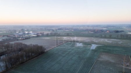This aerial photograph captures the stillness of a misty morning over a rural landscape. The early light diffuses through the haze, casting a gentle glow over the frost-covered fields. In the