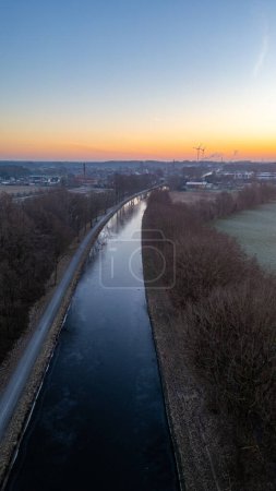 This vertical composition captures the serene beauty of a canal at dawn during the winter season. The canal stretches forward, dividing the landscape, its surface partly frozen and reflecting the
