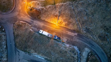 This aerial image captures a lone truck as it traverses a winding road at dusk. The scene is illuminated by a warm streetlight that casts a golden hue on the tarmac, contrasting with the cool tones of