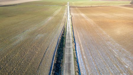Photo for This image offers an aerial view of a long country road stretching into the horizon, dividing two expansive fields. The left field exhibits rows of crops that have been harvested, leaving behind - Royalty Free Image