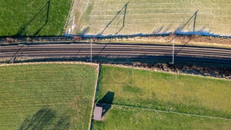 Dieses Bild bietet eine Luftaufnahme von Eisenbahngleisen, die landwirtschaftliche Felder durchschneiden. Die Bahnen erzeugen ein starkes lineares Muster, das parallel zueinander verläuft und den Blick in Richtung