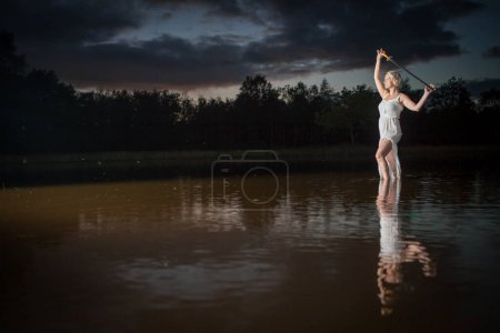 The image depicts an ethereal scene with a young woman in a light, flowy dress standing in a lake at twilight. She is holding a lit sparkler above her head, creating a dynamic contrast with the dusky