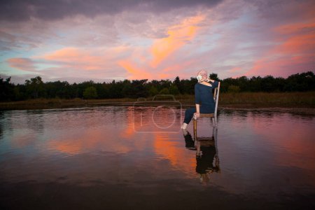 The image captures a serene scene featuring a single individual standing in calm waters at sunset. The person is viewed from behind, creating an air of mystery and contemplation. They are wearing a