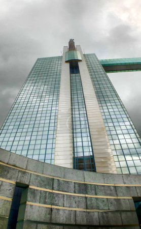 The image captures the imposing facade of a modern skyscraper as it towers into a brooding stormy sky. The buildings glass surface reflects the moody overcast weather, giving it a mirror-like