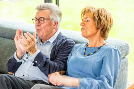 This heartwarming image captures an older couple sharing a joyful moment together on a cozy sofa. The gentleman, clad in a smart casual shirt and glasses, is animatedly clapping his hands with a broad