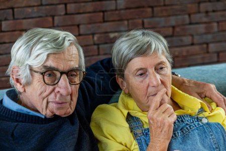 This image provides a candid capture of a senior couple in a moment of reflection. Seated comfortably against a brick wall backdrop that adds a touch of rustic charm, the gentleman is dressed in a