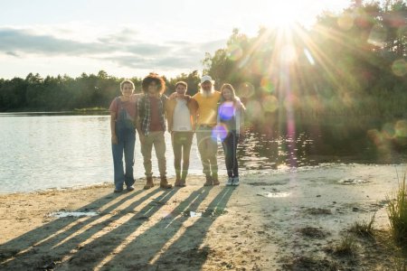 This photo captures a warm moment among a group of friends enjoying the golden hour by a serene lake. The sun, setting in the background, creates a radiant backdrop and casts long shadows on the sand