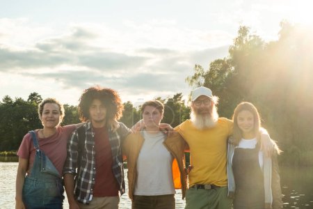 A group of friends stands together by a lake, backlit by the soft, warm light of the setting sun. They represent a range of ages and ethnicities, signifying the beauty of diverse friendships. The glow