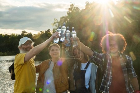 Photo for This image captures a lively moment among a diverse group of friends, raising their water bottles in a toast against the setting suns rays by a calm lake. The sun, positioned between them, creates a - Royalty Free Image