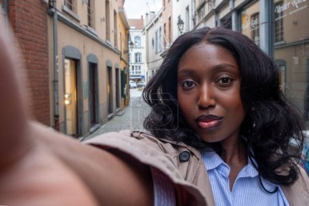 The photograph is a self-portrait of a young African woman extending her arm to capture the photo. She is standing in a narrow city alley, with classic European architecture and cobbled streets in the