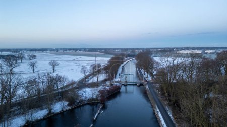 The aerial image captures the serene flow of a canal as it cuts through a vast expanse of snow-covered fields at dusk. The canal, flanked by roads and rows of bare trees, acts as a lifeline in the