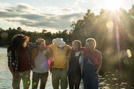This image depicts a group of young adults in a relaxed and intimate embrace, enjoying the company of each other by a tranquil lake at sunset. The suns rays create a radiant halo around the group