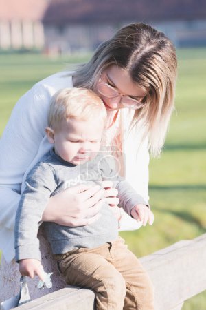 Photo for A young Caucasian woman is seen comforting a toddler in her care, as they sit on a park bench in the early morning light. The woman leans in closely, a gesture of protection and reassurance, as the - Royalty Free Image
