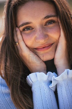 IThis is an intimate close-up portrait of a young woman in natural light. She rests her face in her hands, her elbows seemingly on a surface out of view, and her eyes hold a reflective gaze. The warm