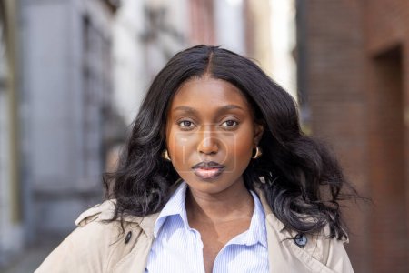 This portrait captures a young African American black woman with a confident gaze standing in an urban alleyway. Her stylish attire, consisting of a classic trench coat over a blue-striped shirt