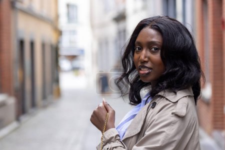 This engaging portrait captures a stylish young African woman turning to give a glance over her shoulder on a quiet city street. Her fashion sense is on display, combining a classic trench coat with a
