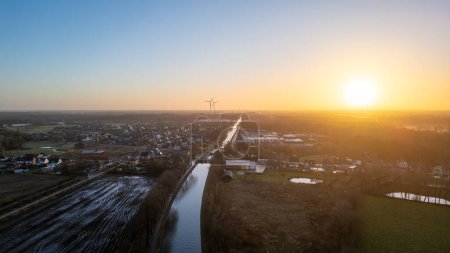 This is an aerial shot captured during sunset, casting a warm glow over a small rural town. The view encapsulates a blend of natural and man-made landscapes, with fields and buildings stretching