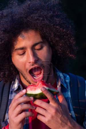 This image features a man of North African origin captured in the midst of relishing a bite from a succulent slice of watermelon. He sports a voluminous, curly afro, and his facial expression is
