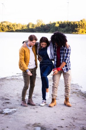 Photo for This poignant image depicts a moment of care and concern among friends at a lakeside setting. A young Asian woman in the center appears to have an injured foot, which she is holding with both hands - Royalty Free Image