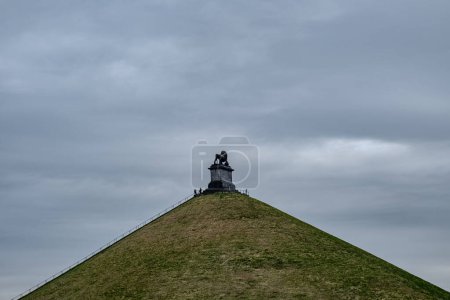 Dieses Foto zeigt einen düsteren Blick auf Besucher, die den Löwenhügel besteigen, das prominente Denkmal am Ort der Schlacht von Waterloo. Der wolkenverhangene Himmel und die steilen Hügel führen das Auge