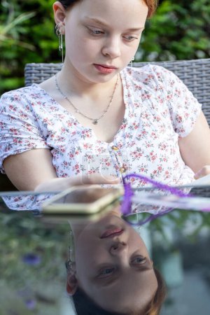 A thoughtful young woman with reddish hair is pictured reading a book, captured from an angle that includes her reflection on a glass surface. The reflection adds a layer of depth, portraying a
