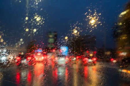 This image captures the blurry lights of a city traffic scene seen through a rain-splattered windshield during nighttime. The droplets on the glass create a soft-focus effect on the red brake lights