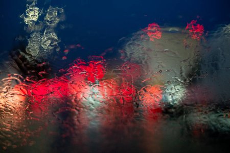 Cette image montre une vue abstraite éblouissante des lampadaires et des phares de véhicules diffusés à travers un pare-brise de voiture trempé de pluie la nuit. La pluie crée une texture fluide et dynamique sur le verre