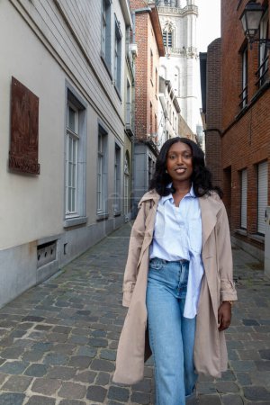 Esta imagen muestra a una joven mujer negra de pie con confianza en medio de un antiguo callejón europeo. El contraste entre su atuendo moderno, incluyendo una camisa blanca crujiente, vaqueros casuales, y un
