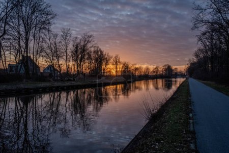 This tranquil image captures the serene beauty of a riverside pathway at twilight. The setting sun casts a warm glow on the horizon, reflected in the still waters of the river, while silhouetted trees