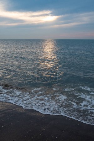 This serene image showcases the quiet beauty of an ocean shoreline at sunset. The suns dwindling light breaks through a gap in the clouds, casting a soft, golden reflection across the gentle waves