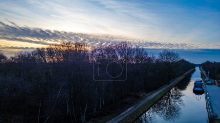 This image captures the serene transition from day to night as twilight descends over a calm canal flanked by bare trees in the winter. The sky, streaked with clouds, is a tapestry of blues and the