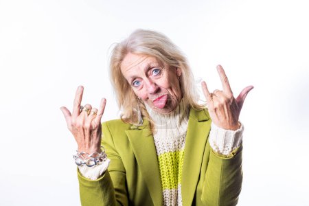 This lively image shows an elderly Caucasian woman with blonde hair making the rock n roll hand sign with both hands. She sticks out her tongue playfully and has a mischievous twinkle in her blue