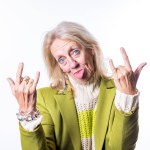 This lively image shows an elderly Caucasian woman with blonde hair making the rock n roll hand sign with both hands. She sticks out her tongue playfully and has a mischievous twinkle in her blue