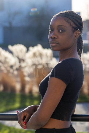 Auf diesem Bild lehnt eine nachdenkliche Afrikanerin an einem Metallgeländer, im Hintergrund ein Stadtpark mit sanftem Fokus. Sie blickt in die Ferne, ihr Gesicht teilweise vom sanften Sonnenlicht erhellt, das