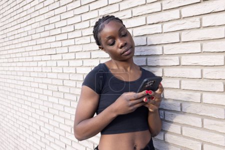 Dieses Bild zeigt eine junge Afrikanerin, die in ihr Smartphone vertieft vor einer weißen Backsteinwand steht. Ihre Haltung ist entspannt, und sie hält das Telefon mit beiden Händen, was darauf hindeutet, dass sie