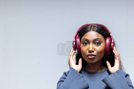 Das Bild zeigt eine afroamerikanische Frau, die einen Moment der Musik genießt, wie die hochroten Kopfhörer beweisen, die sie trägt. Sie hält sie sanft mit beiden Händen, und ihre Augen sind sanft gerichtet