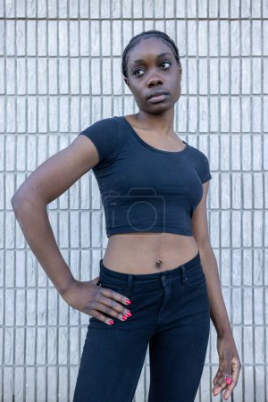 In the image, a poised African woman stands before a white tiled backdrop, her hand casually resting on her hip. Dressed in a black cropped top and high-waisted jeans, she gazes off into the distance