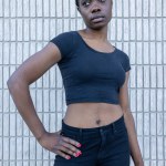 In the image, a poised African woman stands before a white tiled backdrop, her hand casually resting on her hip. Dressed in a black cropped top and high-waisted jeans, she gazes off into the distance
