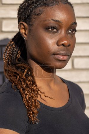 Foto de Este retrato captura a una joven africana mirando a lo lejos, iluminada por la cálida luz del sol que baña un lado de su rostro. Su cabello trenzado, adornado con tonos caramelo, cascadas sobre - Imagen libre de derechos