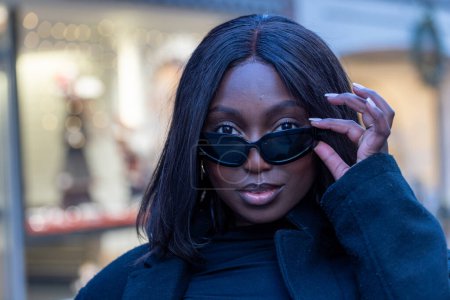 The image presents a moment captured of an African American woman with a playful yet poised expression, as she adjusts her trendy cat-eye sunglasses. Her black turtleneck and coat ensemble speak of