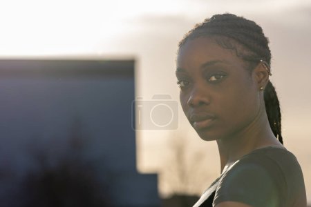This image captures a young African American woman looking calmly towards the camera, her features softened by the diffused light of the setting sun. The urban backdrop is blurred, with the