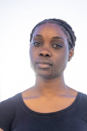 Dies ist ein frontales Porträt einer afrikanischen Frau mit einem gelassenen Gesichtsausdruck vor einem strahlend weißen Hintergrund. Ihre Haare sind zu ordentlichen Zöpfen gestylt, die ihr Gesicht symmetrisch umrahmen. Die sanfte Beleuchtung wäscht