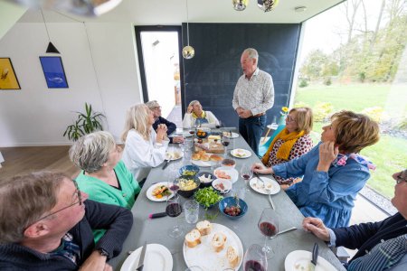 Captured in a modern and airy home setting, this image features a group of senior individuals enjoying a meal together. The host stands, engaging his guests with a story or toast, while the seated