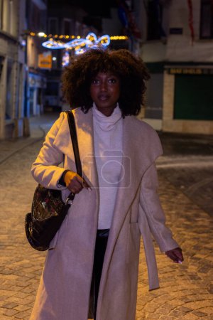 Dieses Bild zeigt eine modische Frau, die nachts auf einer Kopfsteinpflasterstraße spaziert, angestrahlt vom Ambiente des Lichterglanzes der Stadt und der festlichen Dekoration. Ihre selbstbewusste Haltung und ihr direkter Blick